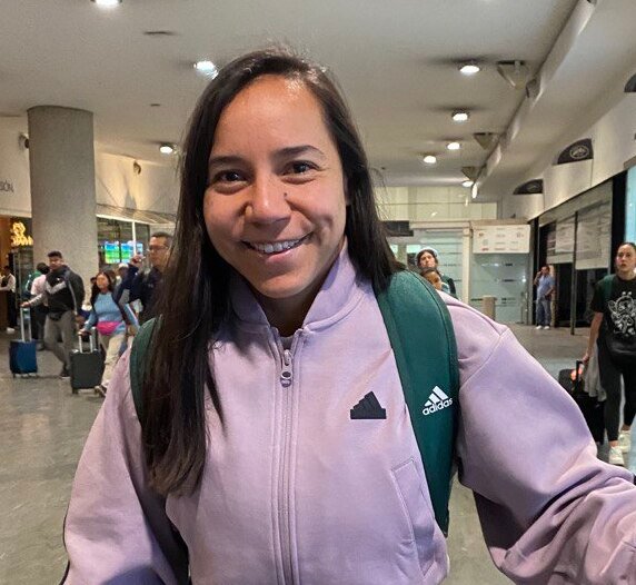 Charlyn Corral sonríe con una sudadera morada y una mochila en un aeropuerto